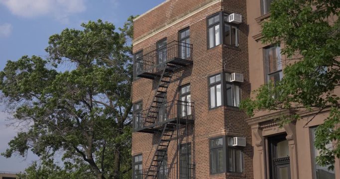 A daytime exterior establishing shot of a typical brick apartment building with fire escapes in Brooklyn.  	