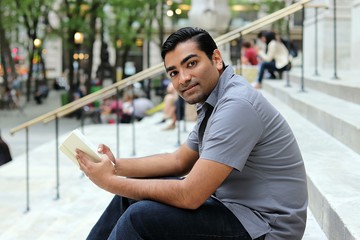 young man holding book and looking out while sitting on urban city steps