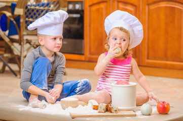 Two siblings - boy and girl - in chef's hats sitting on the kitchen floor soiled with flour, playing with food, making mess and having fun