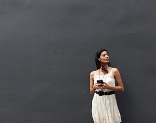 Beautiful brunette female model cell phone looking up while listening to music in headphones in white dress and black belt while standing against grey dark background