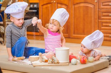 Two siblings - boy and girl - and a newborn kid with them in chef's hats sitting on the kitchen floor soiled with flour, playing with food, making mess and having fun