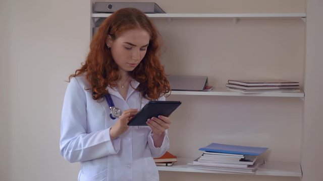 Caucasian doctor with red curly hair reading medical records using digital tablet. Candid adult woman wearing in white lab coat and phonendoscope standing in hospital.