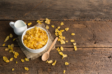 white bowl of cornflakes on sackcloth with wooden spoon and little pitcher of milk on wooden background with copy space ,still life , in concept breakfast
