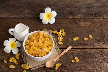 Obraz na płótnie Canvas white bowl of cornflakes on sackcloth with wooden spoon and little pitcher of milk and beautiful flower on wooden background with copy space ,still life , in concept breakfast