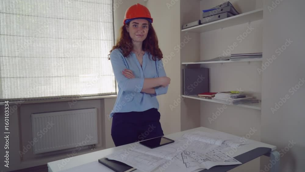 Poster Redheaded engineer at work using digital tablet. Professional female looking at the camera with arms crossed. On the desk sketch with technical drawing some building.