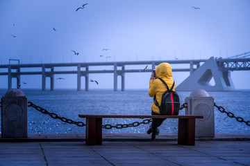 rear view of a female tourist sitting on bench while taking a picture of Dalian bay bridge,China.