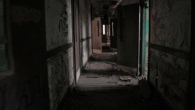 FPV, CLOSE UP: Walking along dark narrow hallway in abandoned decaying psychiatric hospital. Moving past the weathered wooden doors and broken crumbling walls in sinister sanatorium looking for escape