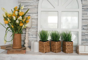 White wooden shelf with bouquet of tulips, grass in pot, greenery in vase, books and candle against stone wall with decorative window. Сlose-up decoration in living room at home