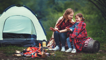 Family mother and child daughter warm their hands by bonfire on camping trip with tent