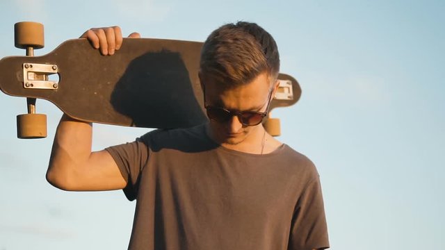 Hipster teenager wears sunglasses holding longboard on his shoulder against sunset, 120FPS slowmotion