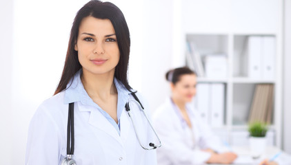 Fototapeta premium Brunette female doctor on the background of colleagues talking to each other in hospital. Physician is ready to help patients. Medicine and health care concept