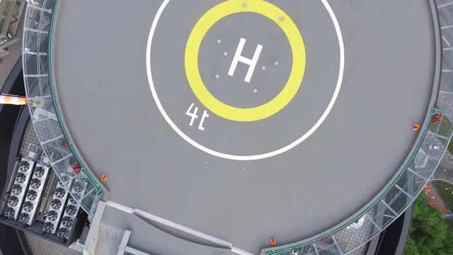 The helicopter is slowly flying over the modern business center on the roof of a group of air conditioners, a helicopter landing pad with a yellow circle, and below is a road with cars and a green