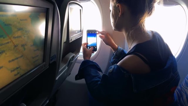 The girl climbs into the porthole and takes pictures of the landscape in the window on the phone. A young girl is traveling by plane. Slow motion 1080p
