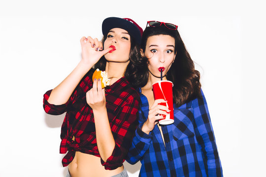 Young hipster girls having fun drinking a soda from straw and holding a burger, happy smile and laugh on the white background.