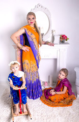 Young blonde woman in a bright Indian saris and jewelery stands next to a white fireplace, and her little daughter sitting together on the floor and wooden horse