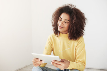 Fototapeta premium Beautiful african girl smiling with closed eyes listening to music in headphones enjoying holding tablet.
