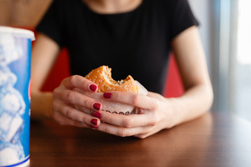 A girl holds a hamburger in a cafe on the background of the drink on the wooden table in fast food dining