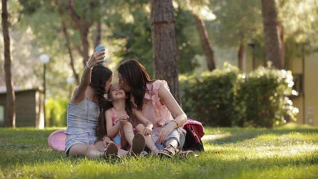 Mom with two daughters in the nature sitting on the grass and making a selfie with a phone