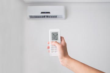 Hand holding remote control, adjusting temperature of air conditioner mounted on a white wall. Indooor comfort temperature