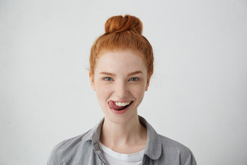 Young teenage female with ginger hair knot sticking out her tongue having funny look isolated over white background. Freckled student girl having fun being happy to finish her studying successfully © WHstudio Leushin N
