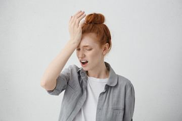 Tired red-haired student female with hair knot having headache after stress on exam. Hipster ginger woman with sorrorful look holding her hand on head trying to find way out in difficult situation