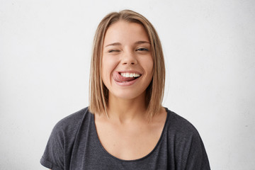 Horizontal portrait of optimistic funny girl with bob hairstyle showing her tongue and blinking eyes while posing against white studio background. Emotional positive young female making grimace