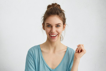 Excited smiling young female with hair knot expressing positive emotions while posing against white background. Natural woman with pleasant wide smile wearing blue loose shirt raising her hand up