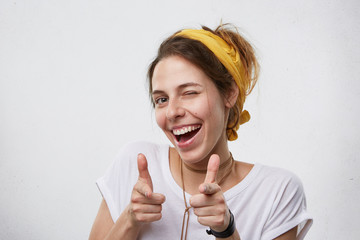 Positive cheerful young woman wearing yellow scarf on head and white casual T-shirt blinking her eyes and smiling pointing at camera with index fingers. Happy attractive woman pointing at you