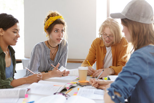 Four mixed race students dicussing something while sitting with books indoors deciding how to make project. European woman having annoyed expreesion while trying to expain material to her groupmate