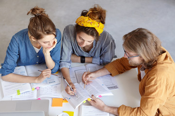 Two pupils working with their tutor doing home assigment sitting at white table. Three college students working with books underline important information on pages having deep look. Scientific work