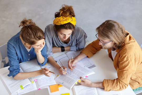 Group of pupils graduating from school preparing for their final exams meeting together learning material. Three friends having conentrated look in book pointing at it with pencils sitting indoor