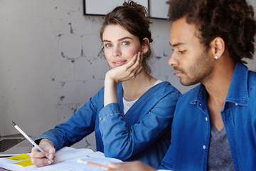 Young attractive teacher teahing her mixed-race African student explaining her theory giving some examples writing rules. Dark-skinned male listening carefully to his teacher having look in books
