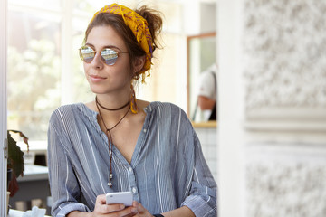 Trendy young woman in shades and yellow headband leaning at table holding mobile telephone standing...