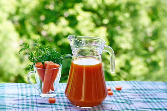 Glass jug of carrot juice and carrots in a glass on a background of green nature.