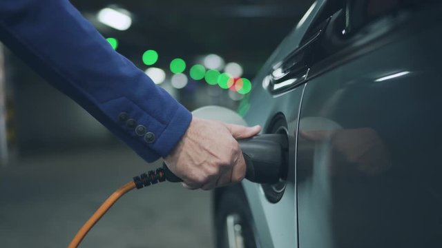 Senior is plugging in power cord to an electric car. Close up. 4K