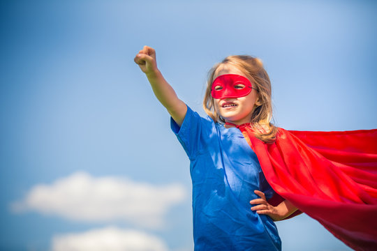 Funny little girl playing power super hero over blue sky background. Superhero concept.