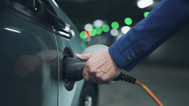 Senior is plugging in power cord to an electric car. Close up. 4K