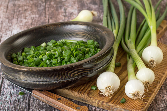  Fresh greenery. Young green onion on a kitchen cutting board and sliced in a wooden bowl on an old table.
