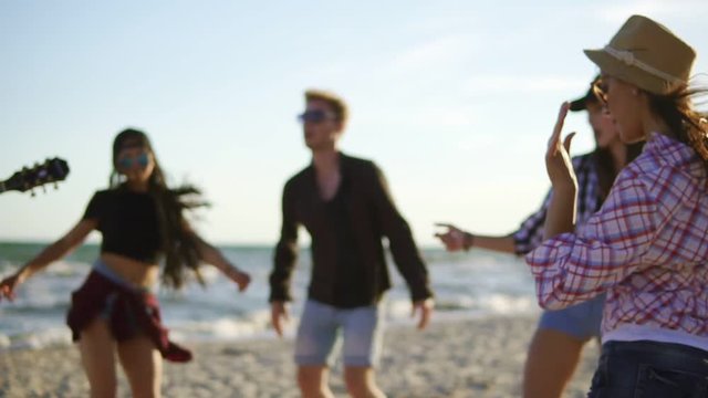 Summer party on the beach. Young friends drinking cocktails, dancing in the cirkle, playing guitar, singing songs and clapping on a beach at the water's edge. Slowmotion shot