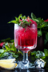 Drink me! Glass of alcohol cold red cocktail decorated with ice, fruit, flowers and mint staying on table in bar. Fresh juice on black background with lemon, strawberry lime.