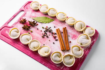 Dumplings home cooking in a bright pink board with flour and seasonings. Colorful board with dumplings, spice - cinnamon sticks, anise and pepper, food, white background