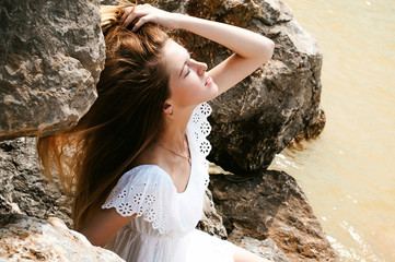 Portrait of slender young woman on stones near the sea. Beautiful girl is resting on coast, enjoying outdoor recreation