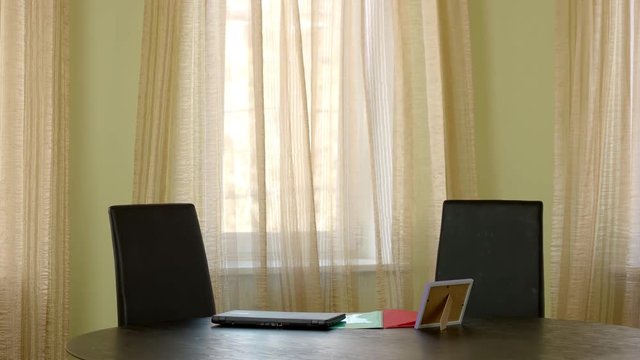 Table with laptop near window. Curtains moving in the wind. Simple office room design.