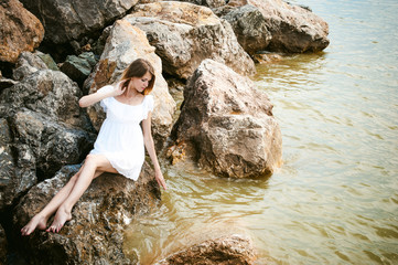 Portrait of slender woman on stones near the sea. Beautiful girl is resting on coast, enjoying outdoor recreation. sexy white dress, graceful long thin tender hands and feet Splashing water