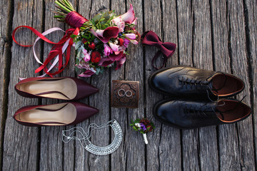 Wedding accessories. Bridal shoes on high heels, jewelry for bride, groom's shoes, bow tie, bouquet of flowers and wedding rings on vintage wooden background