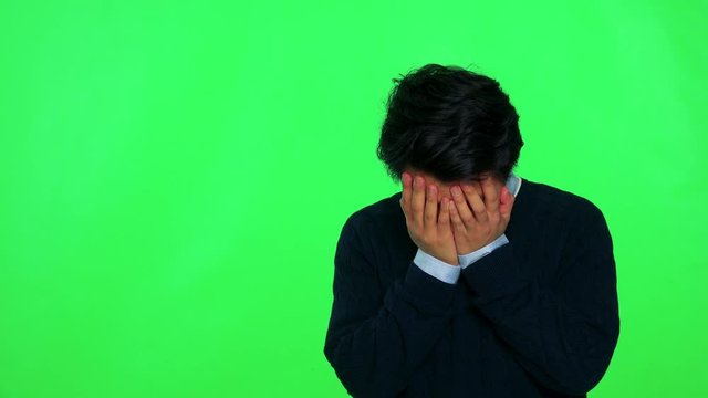 A young Asian man covers his face with his hands, then stops and looks around, confused - green screen studio