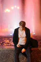 Smiling guy reading message on smartphone, while he working on laptop outside. Young male freelancer working in city street in night time, blurred lights background, free space