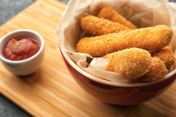 Wooden board with cheese sticks in bowl and tomato sauce on table
