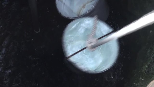 A person is drawing water from a well with a bucket helping with a rope and a pulley. Insert another empty bucket to draw more water. View from above