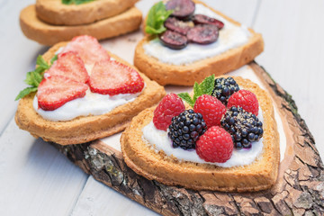 Heart shaped biscuits spread with quark, strawberries, blackberries, raspberries, cherries and a twig of mint presented on a tree disk